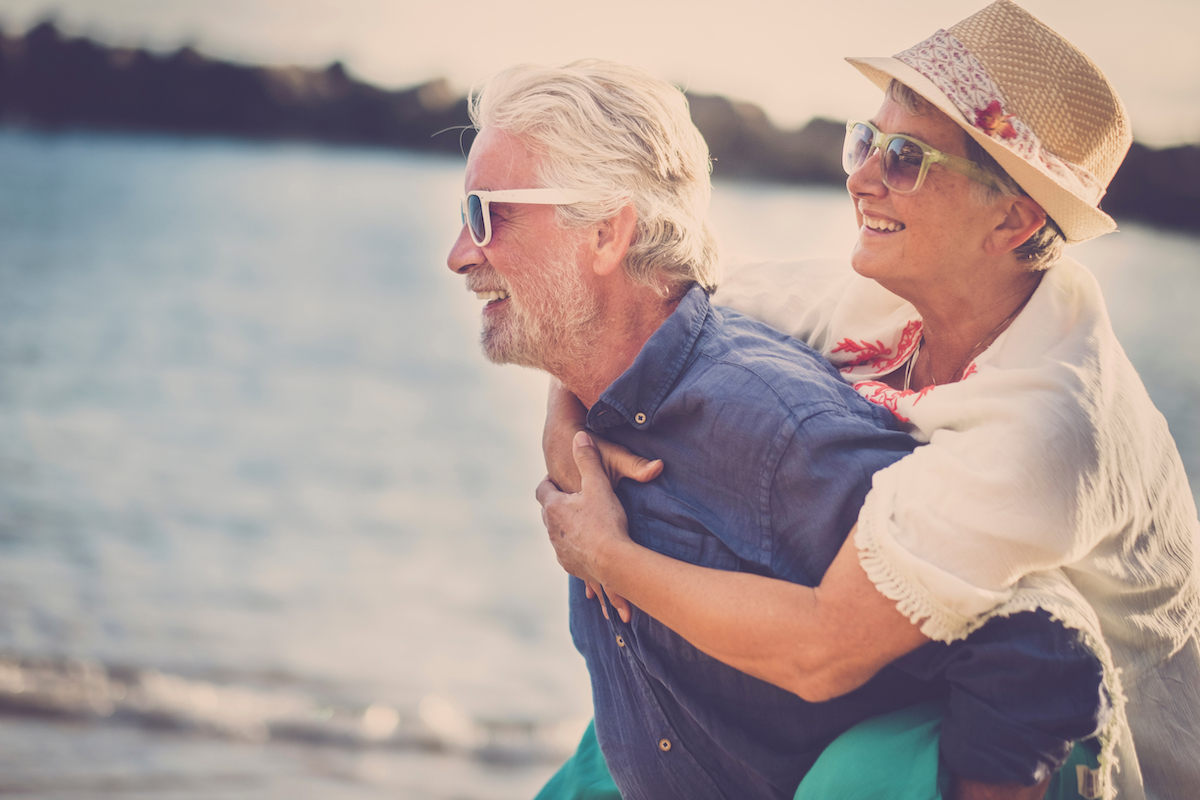 Happy Senior Man Carries Woman on His Back at Beach