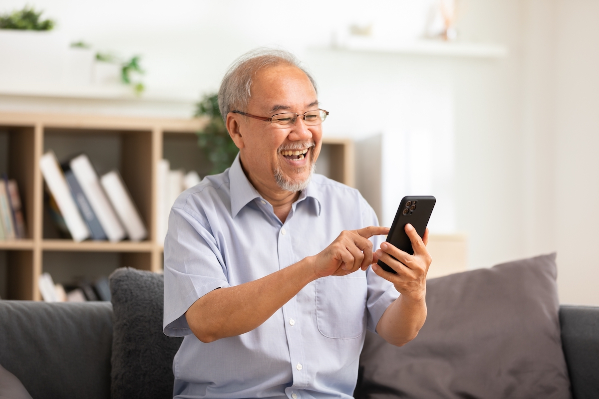 Senior Man Sitting on Couch Smiling and Scrolling Through Phone_Franklin Park® Senior Living