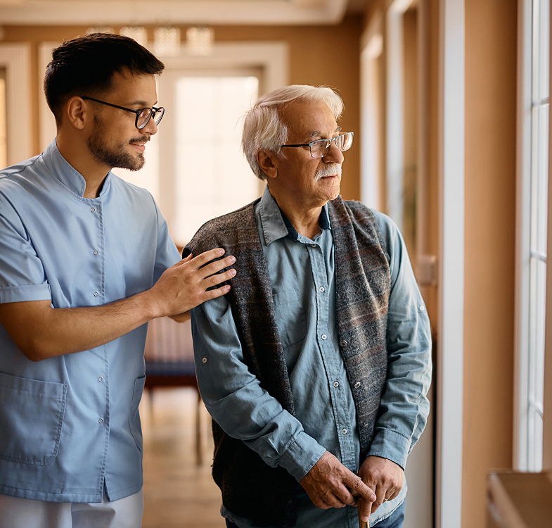 Caregiver supporting elderly man by window indoors